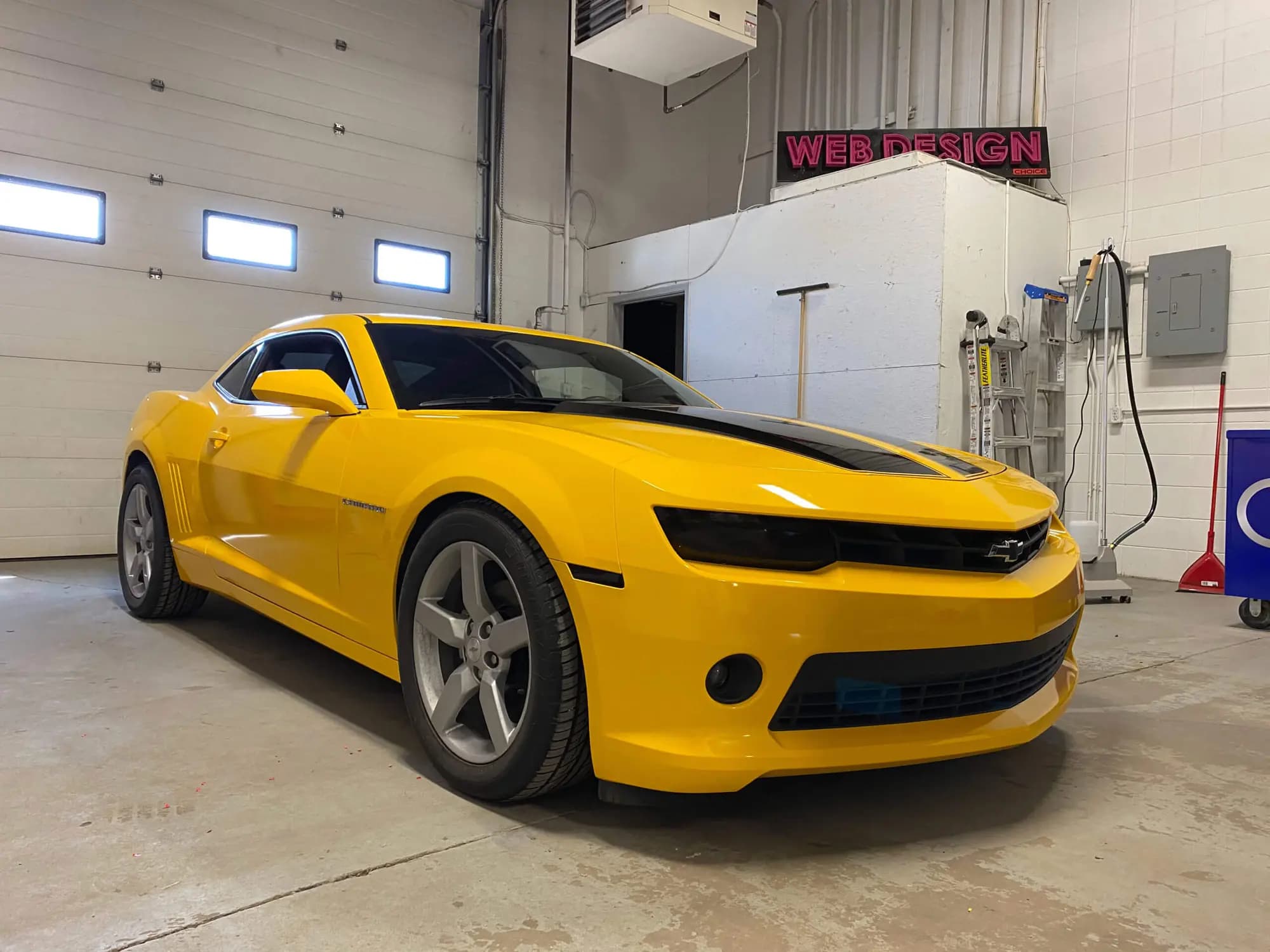 Yellow Chevrolet Camaro coupe with black hood stripes parked in a shop bay, wrapped in Gloss Sunflower