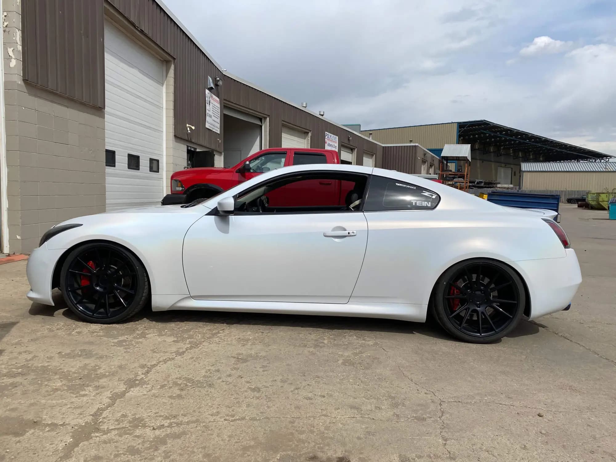 Side profile of a white two-door sports coupe wrapped in Satin Flip Ghost Pearl, on black wheels with red brake calipers, parked outside an industrial shop
