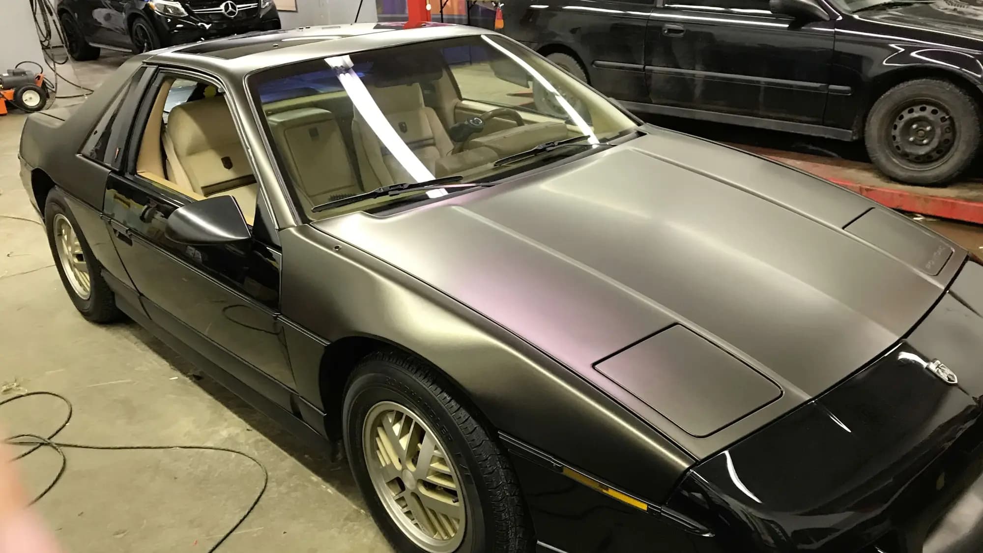 Pontiac Fiero in the shop, its hood, front fenders and roof wrapped in Satin Gold Dust Black beside untouched gloss black bodywork