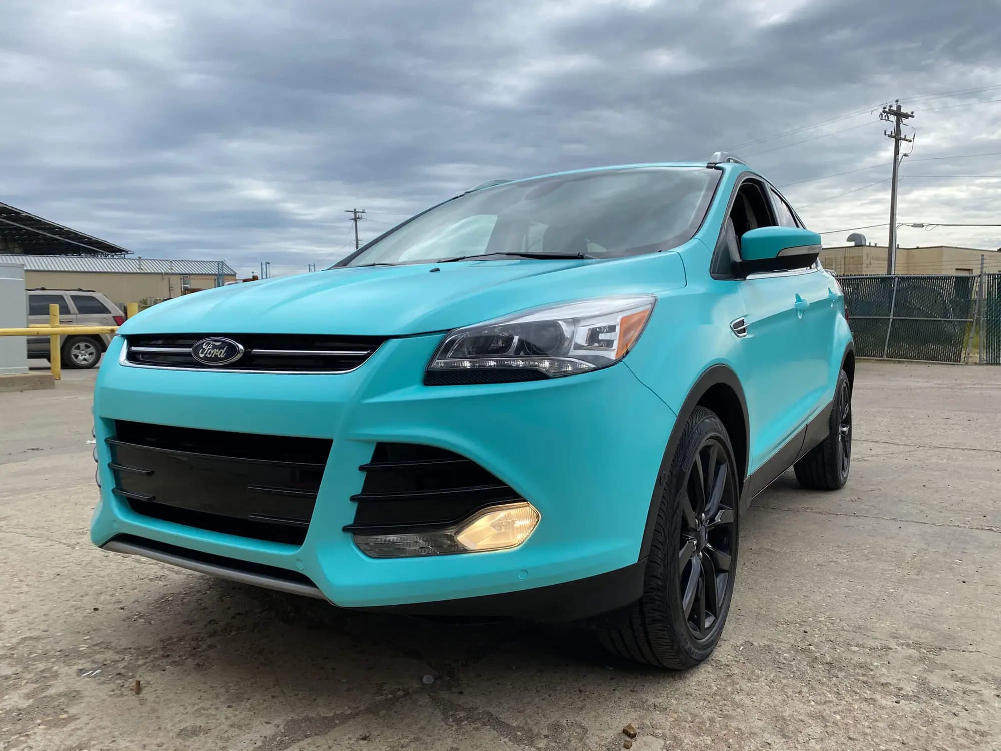 A Ford Escape wrapped in Satin Key West, front three-quarter view in a gravel yard under a grey sky, on black wheels